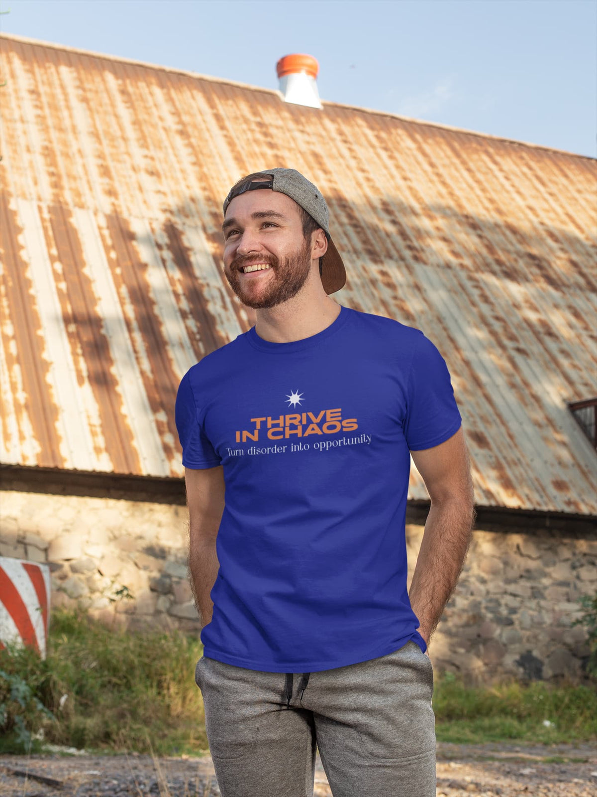 Man wearing a blue t-shirt with 'Thrive in Chaos' text standing in front of a rustic building.