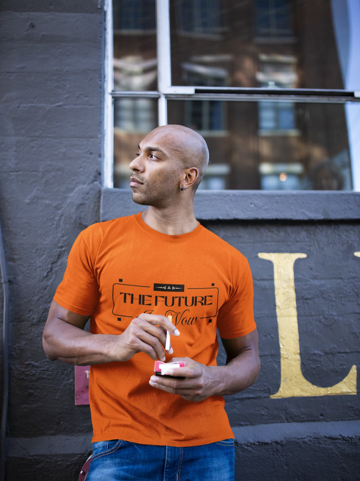 Man wearing an orange t-shirt with text, standing against a dark wall with large letter 'L'.