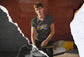 Person wearing a gray t-shirt with a logo, sitting with a skateboard on a textured background