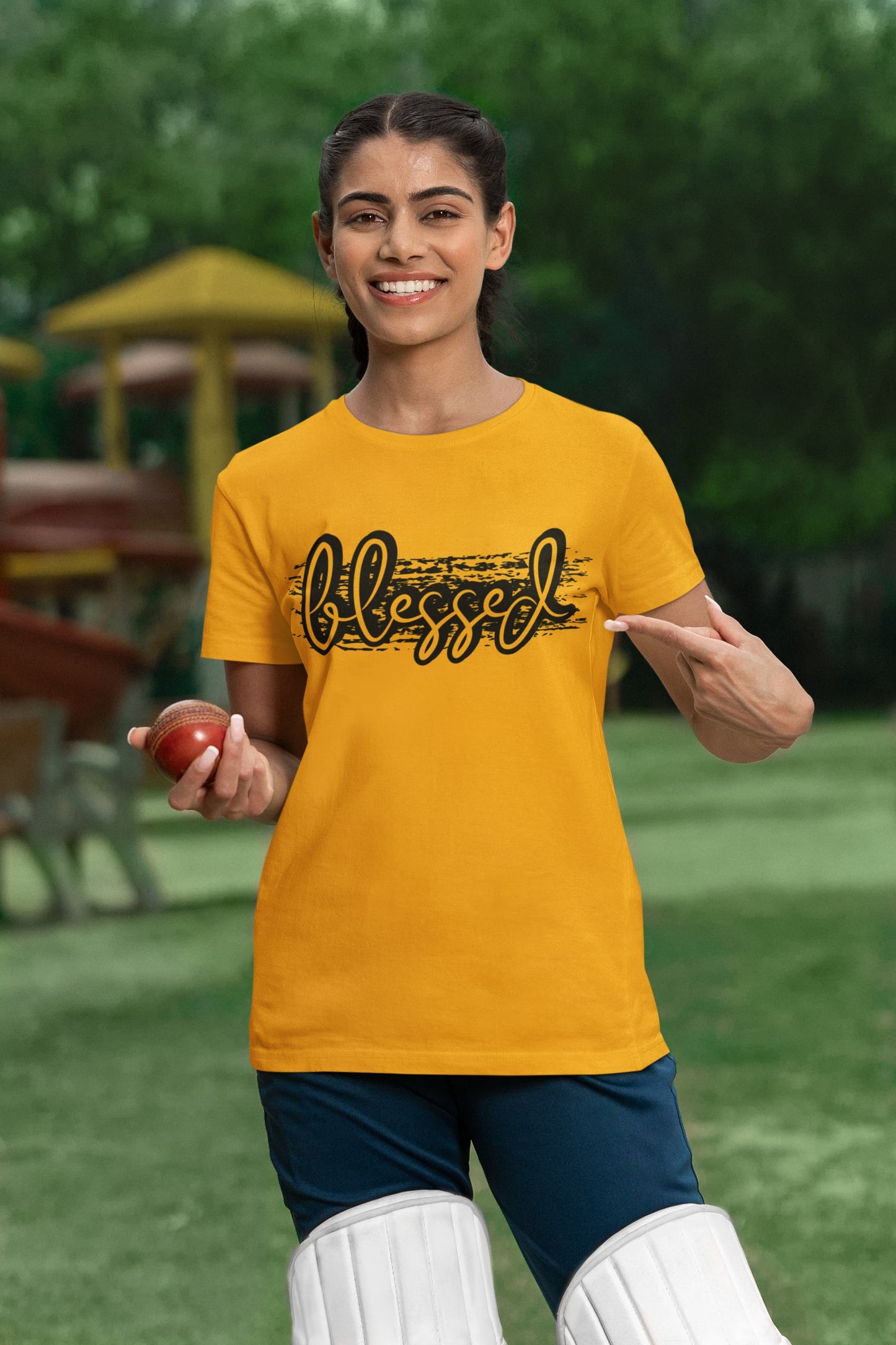 Woman in yellow t-shirt with 'Blessed' text, holding a cricket ball on a grassy field.