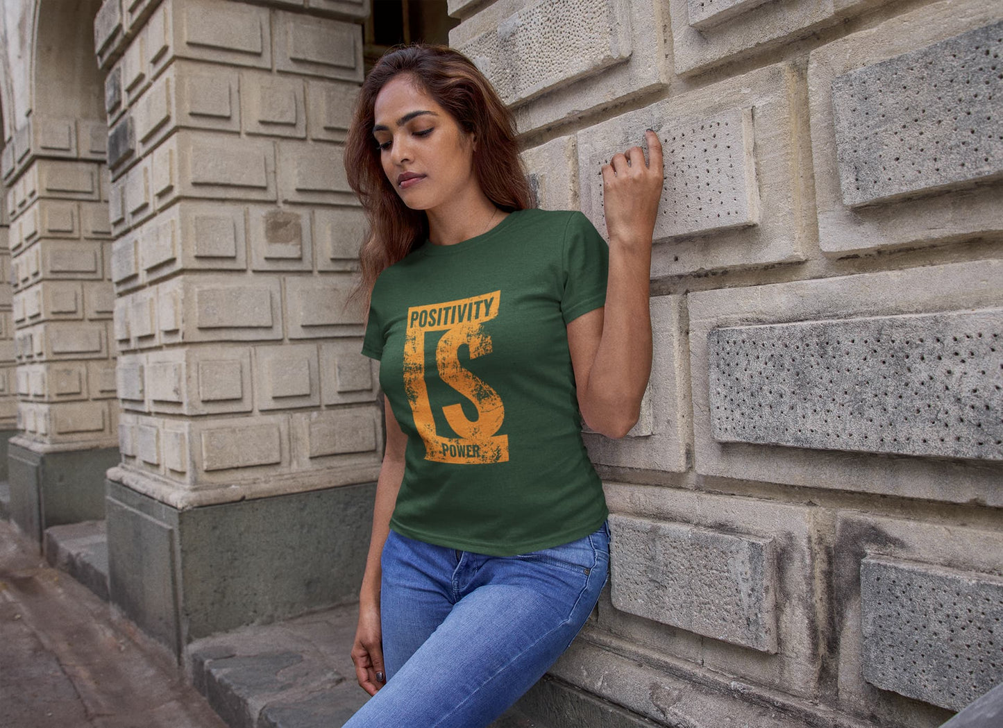 Woman wearing a green t-shirt with 'Positivity' design leaning against a stone wall.