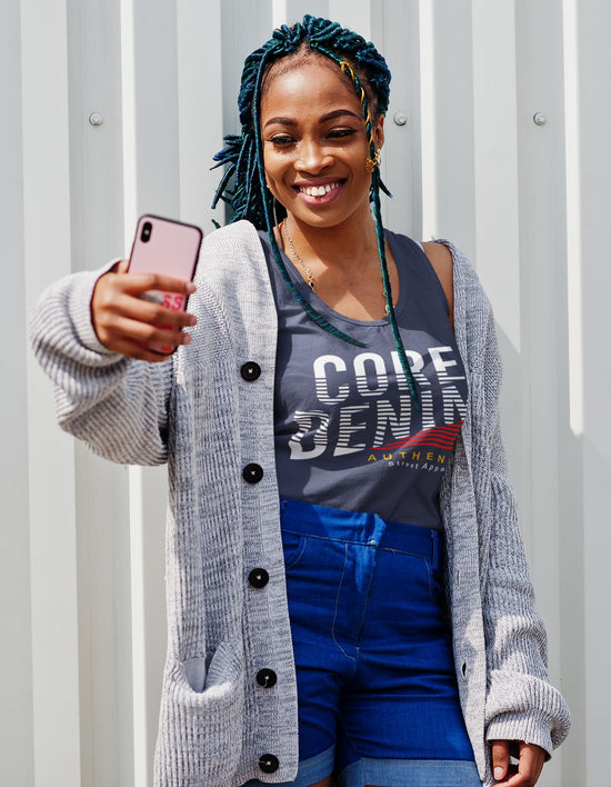 Woman taking a selfie wearing a gray cardigan, gray t-shirt with branding, and blue shorts against a light gray background.