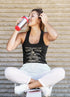 Woman drinking from a red and silver tumbler wearing a black tank top with text.